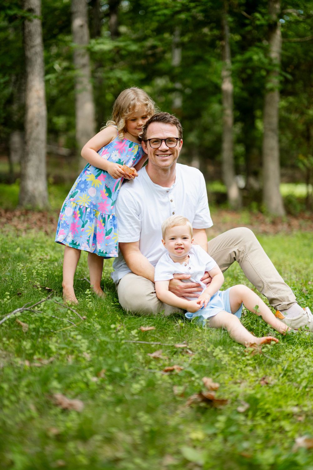 A smiling father sits on green grass with his two young children in a wooded outdoor setting.