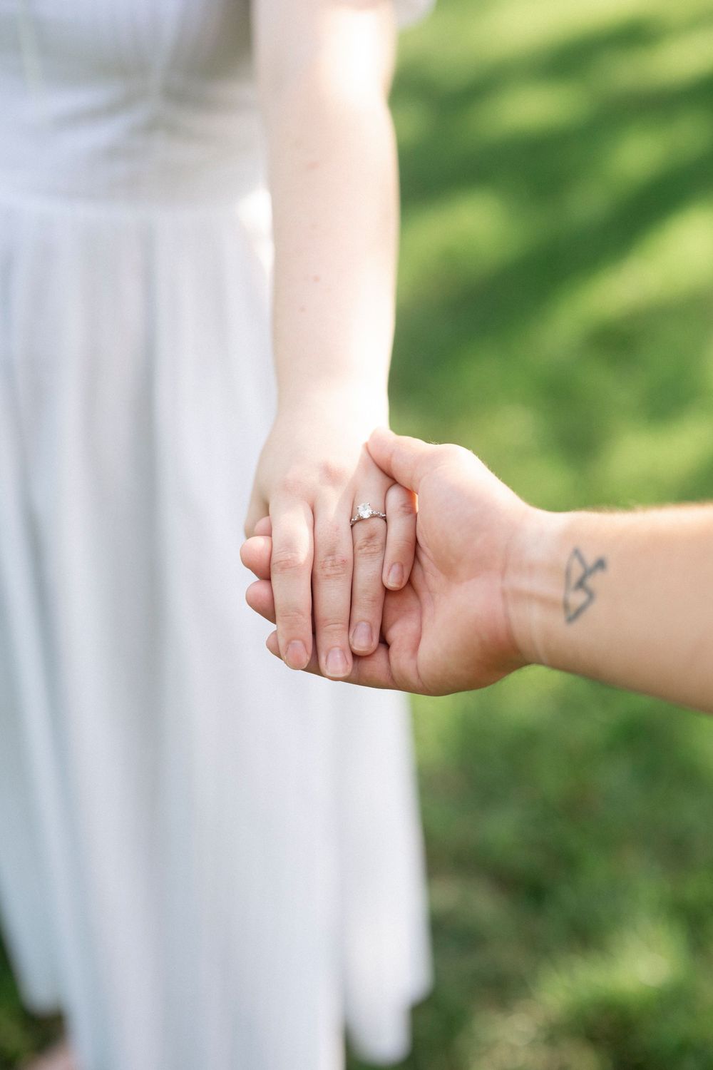 Close-up of couple holding hands outdoors, woman wearing engagement ring in white dress, man with wrist tattoo on green background.