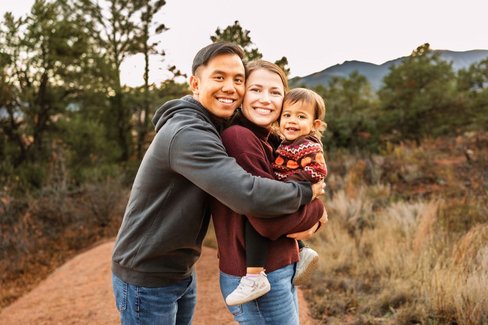 A family embraces outdoors on a mountain trail during golden hour with pine trees and mountains in the background.