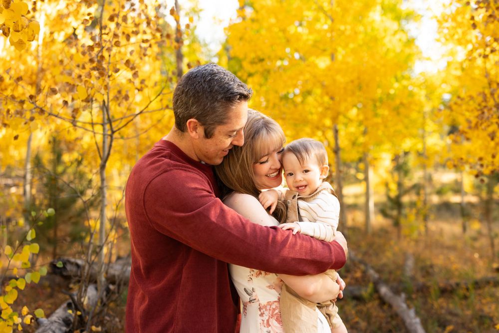 A family shares a tender moment together while standing among vibrant yellow autumn trees in a forest setting.
