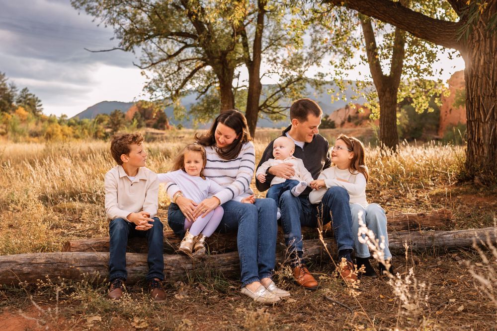 A loving group sits together on a fallen log in a golden autumn field with mountains in the background at sunset.