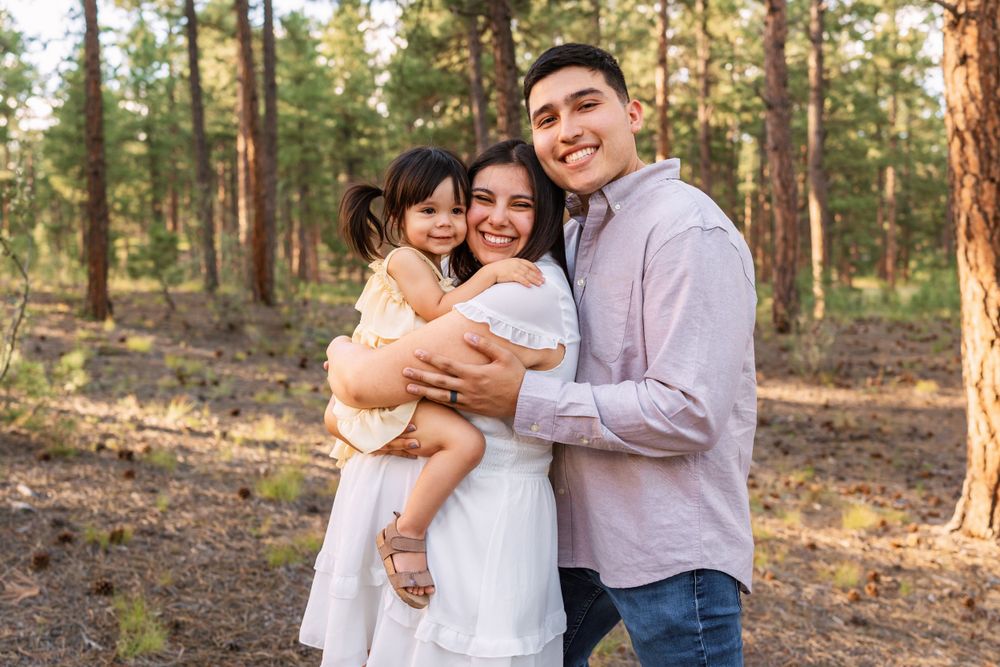 A young family shares an embrace while posing together in a sunlit pine forest setting.
