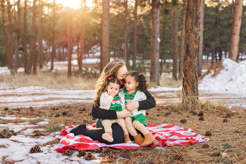 Two people sitting on red plaid blanket in snowy forest during golden sunset amongst pine trees.