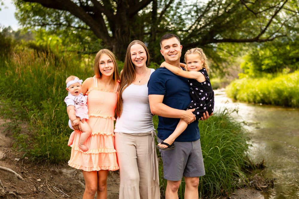 A group of people standing outdoors by a stream on a sunny summer day surrounded by lush greenery.