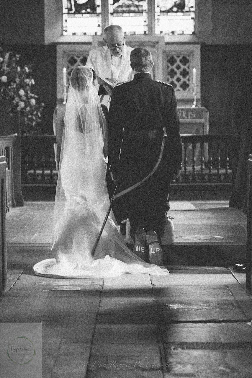 Wedding reportage photo of bride and groom kneeling during church ceremony, groom has written "HELP" onto the soles of his shoes