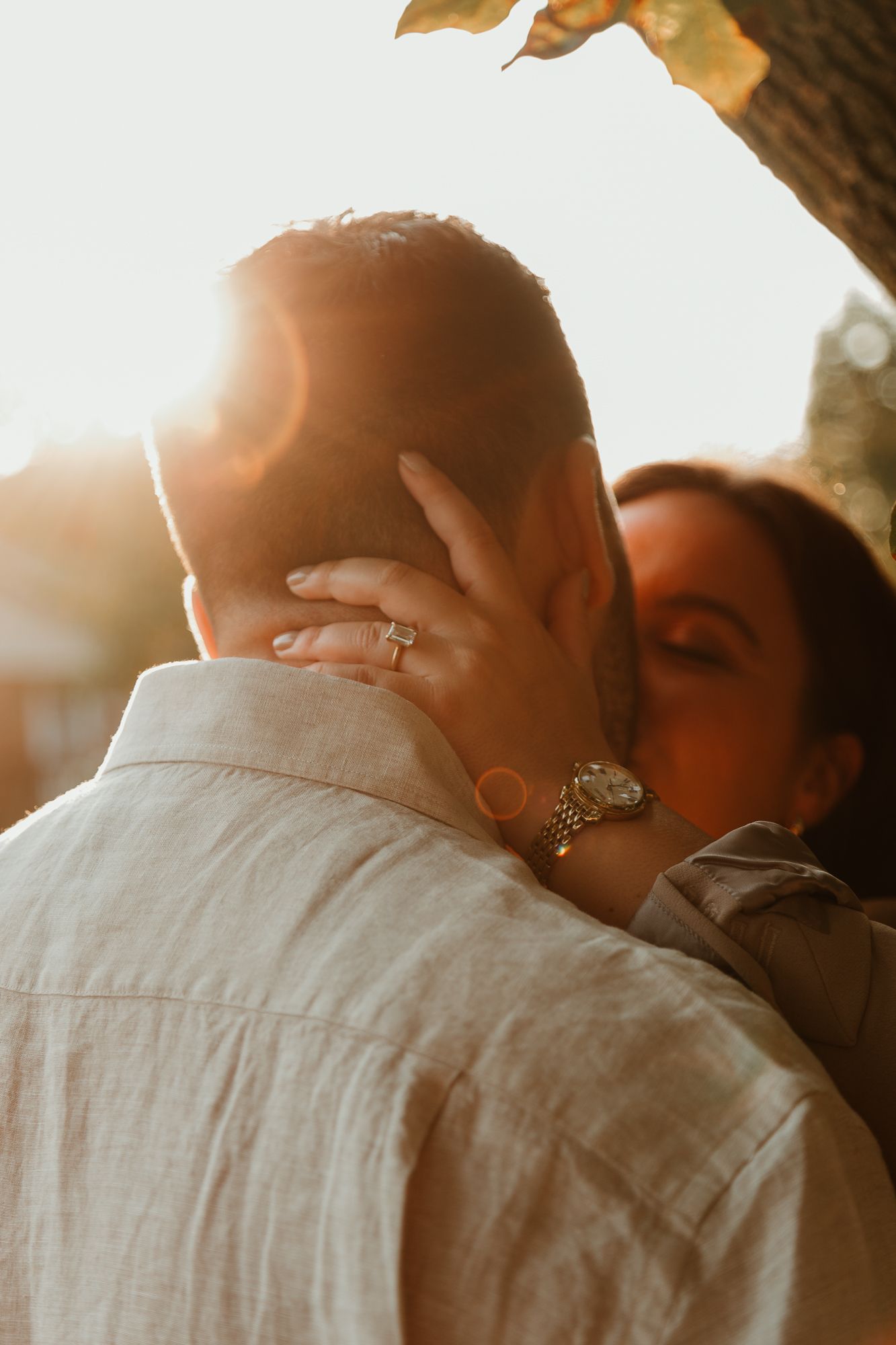 A romantic couple sharing a kiss during sunset, with a warm golden glow illuminating their intimate moment while showing off engagement.
