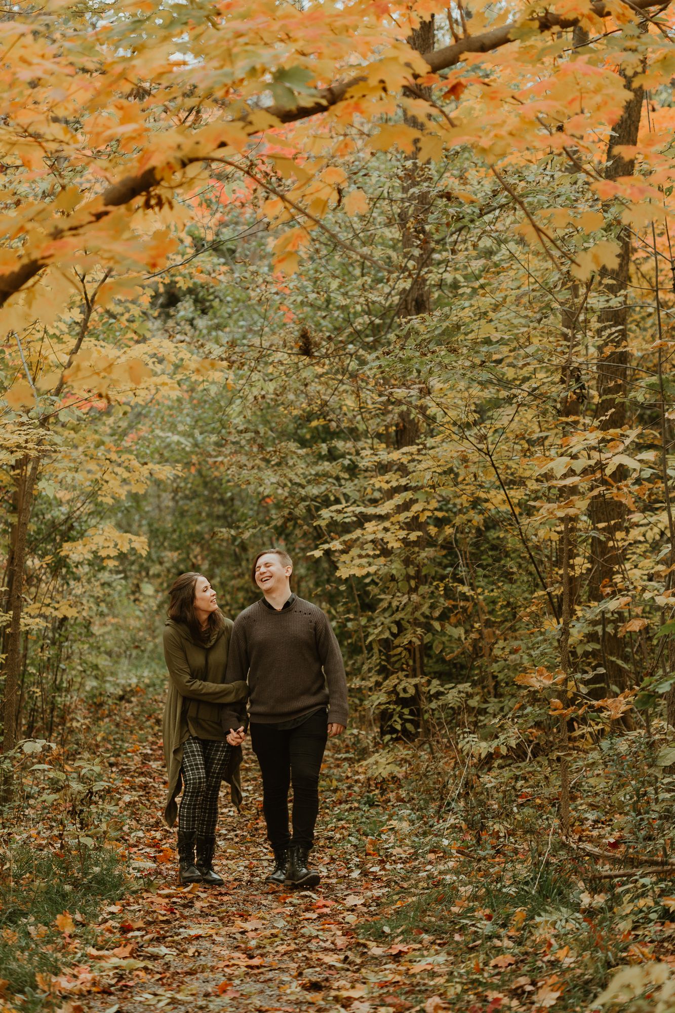 A couple walks hand in hand through a forest path surrounded by vibrant autumn foliage and fallen leaves.