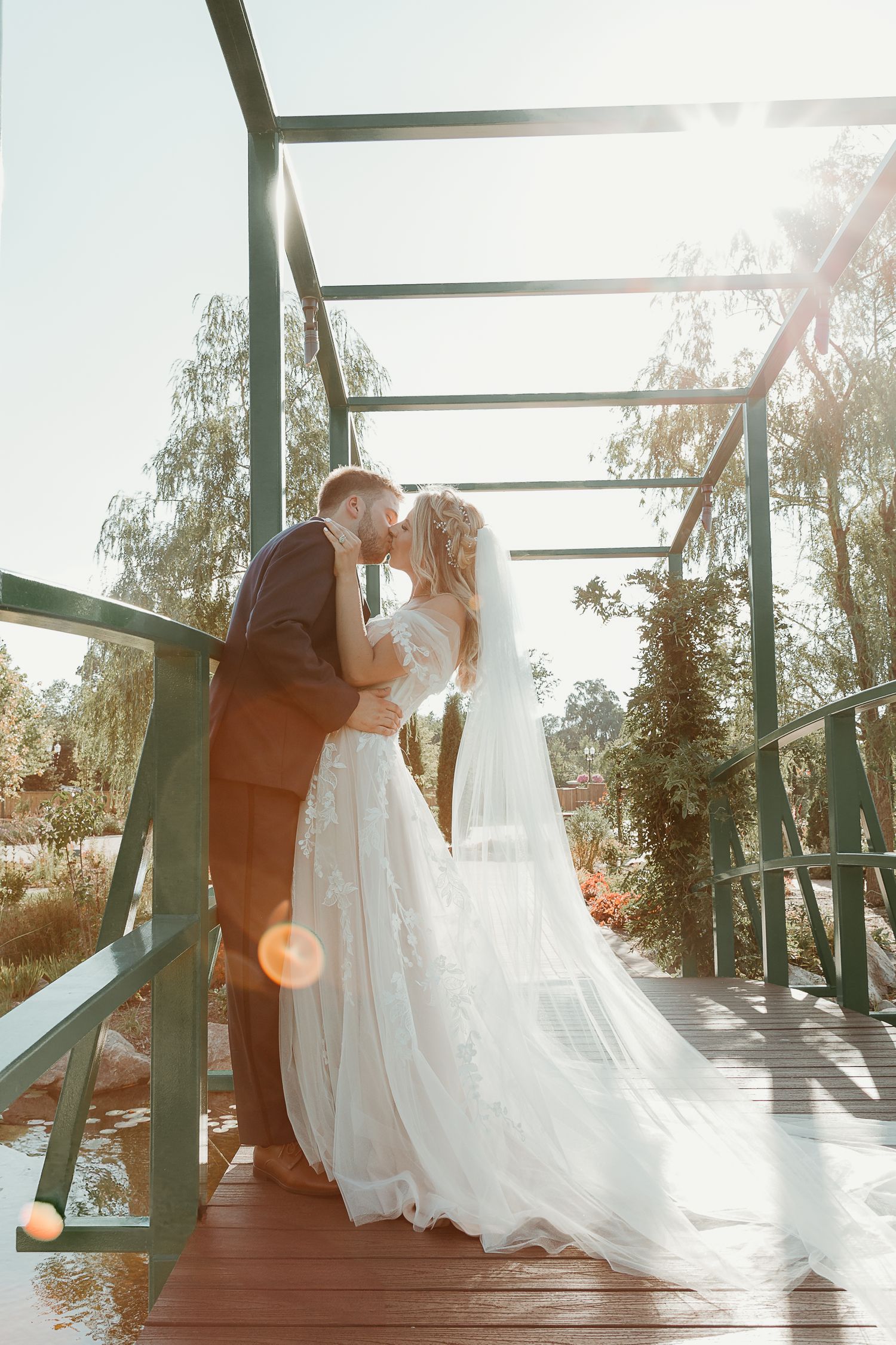 A romantic outdoor wedding moment captured on a wooden deck with sunlight streaming through trees in the background.