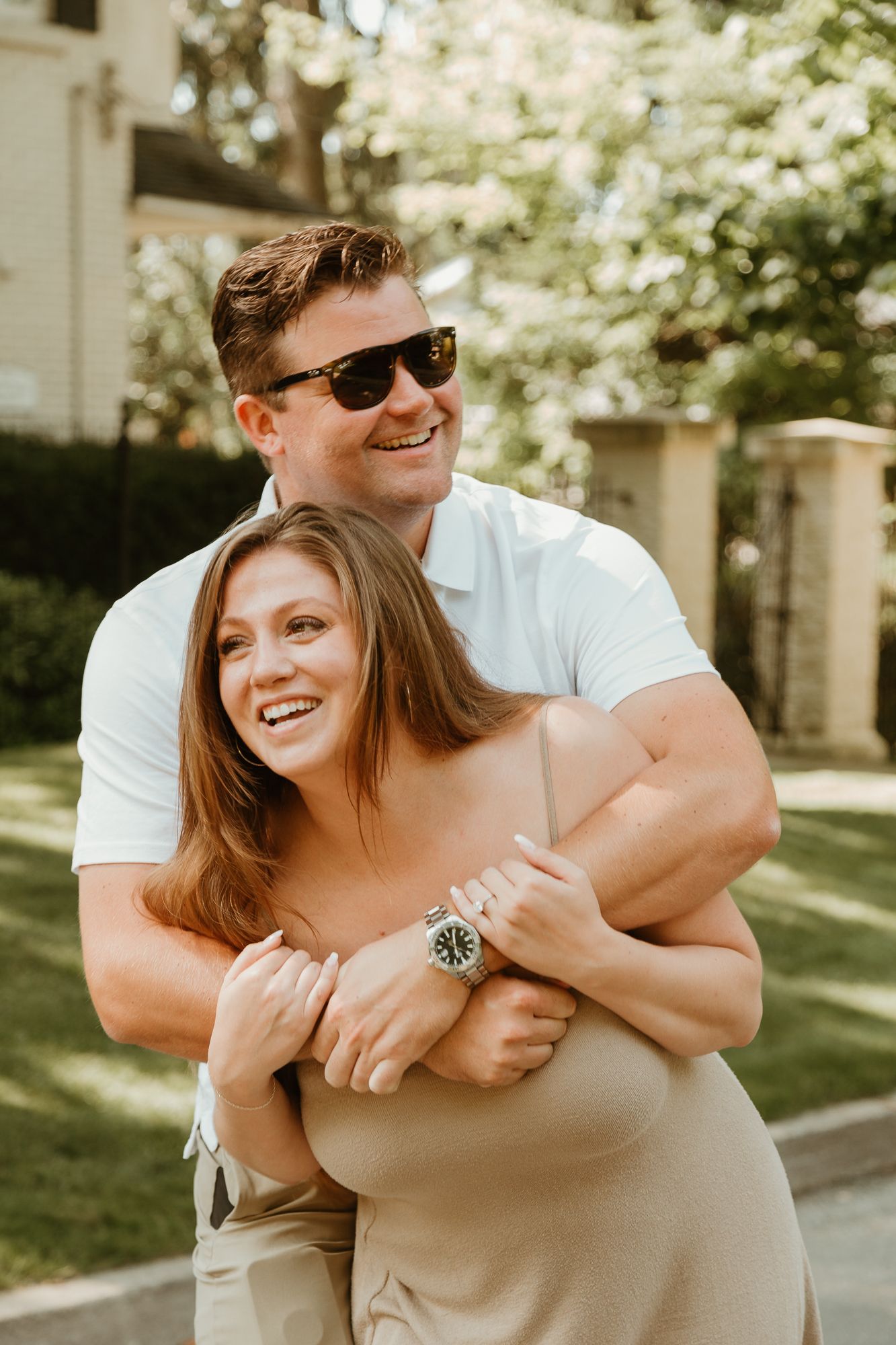 A playful couple embraces and laughs together in a sunny outdoor setting wearing casual summer attire.