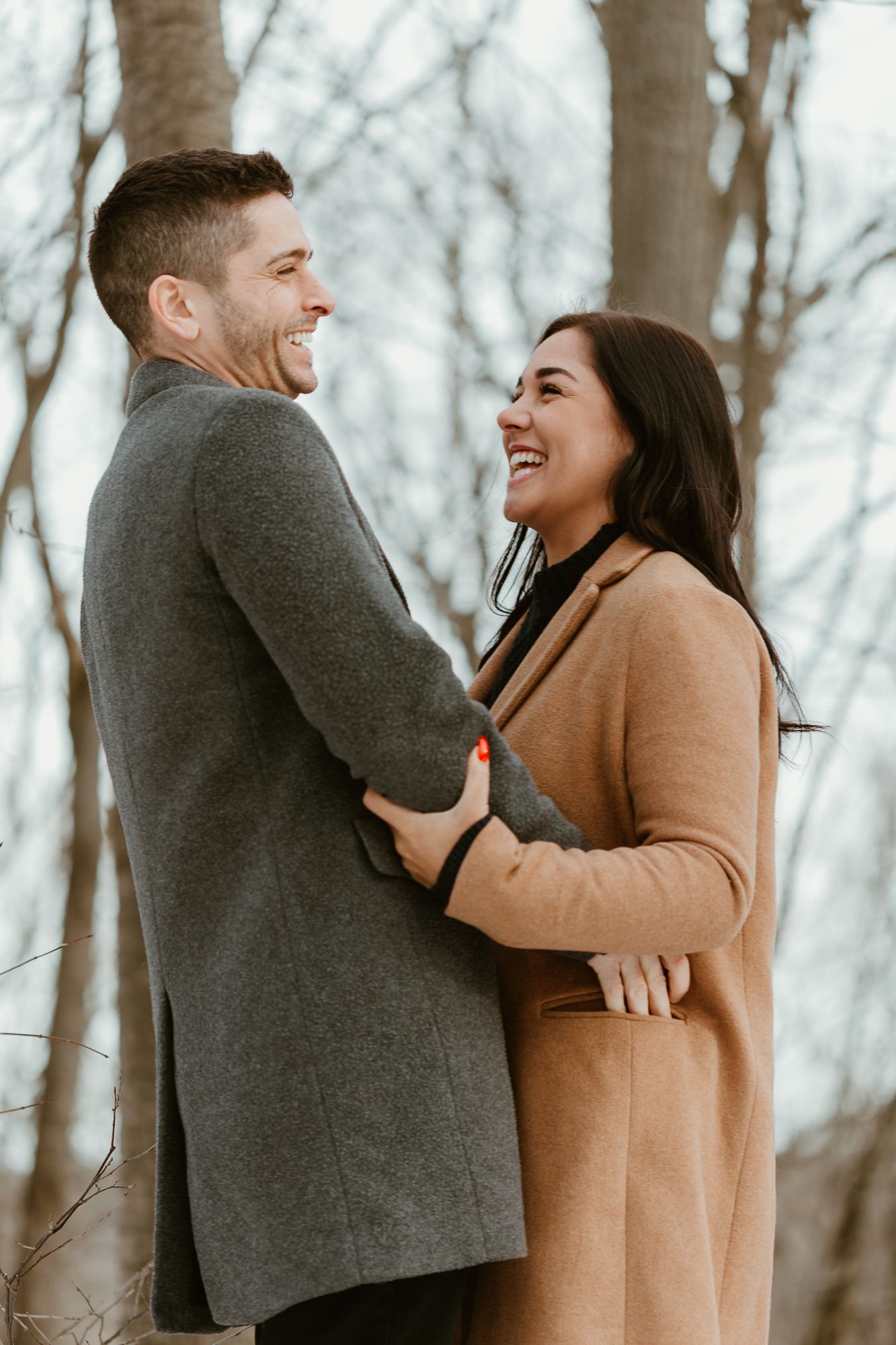 A couple in winter coats share joyful laughter while embracing outdoors against a backdrop of bare trees.