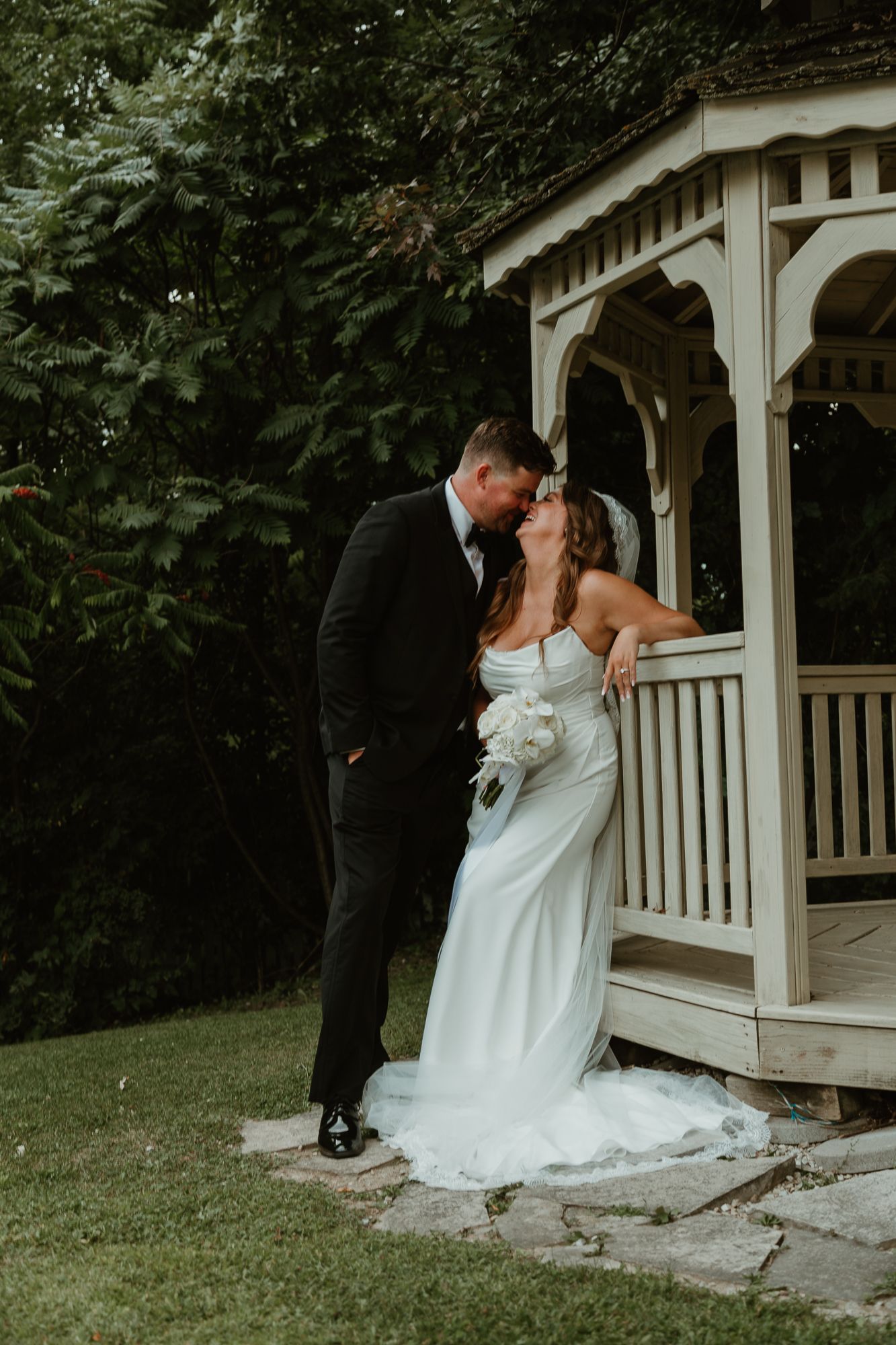 A romantic wedding couple shares an intimate moment in front of a white garden gazebo surrounded by lush greenery.