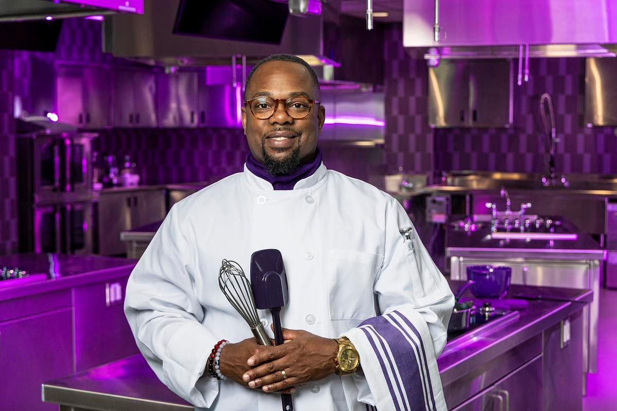 A chef in a white coat stands confidently in a professional kitchen illuminated with purple lighting.
