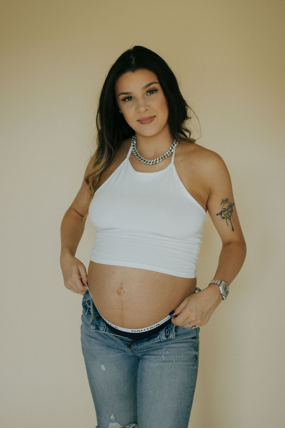 A person poses in a white halter crop top and distressed jeans against a beige wall, showing visible tattoos and jewelry.