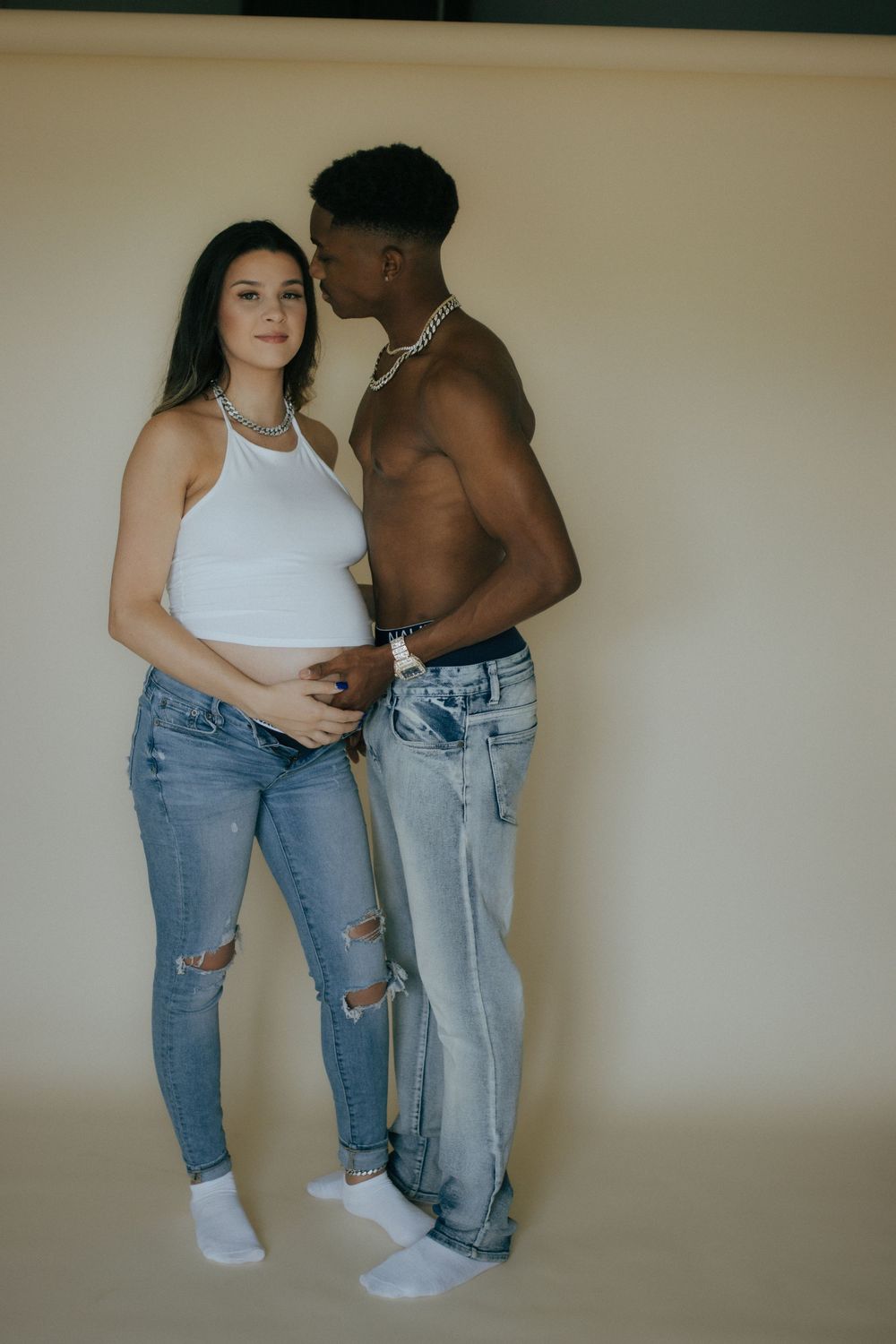 Two people in casual attire pose intimately against beige wall wearing denim jeans and white tops.