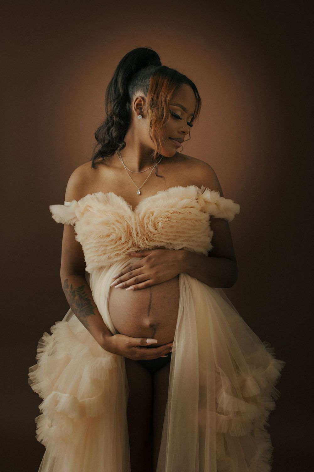 A maternity portrait features an elegant figure in a flowing off-shoulder cream gown against a warm brown backdrop.