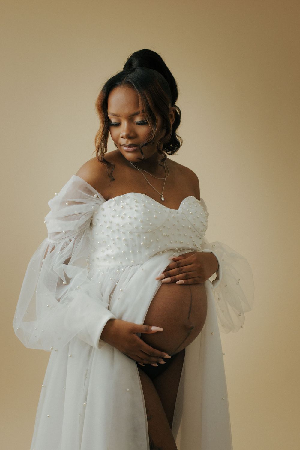 A romantic maternity photo features someone in an ethereal white off-shoulder gown gently cradling their baby bump.