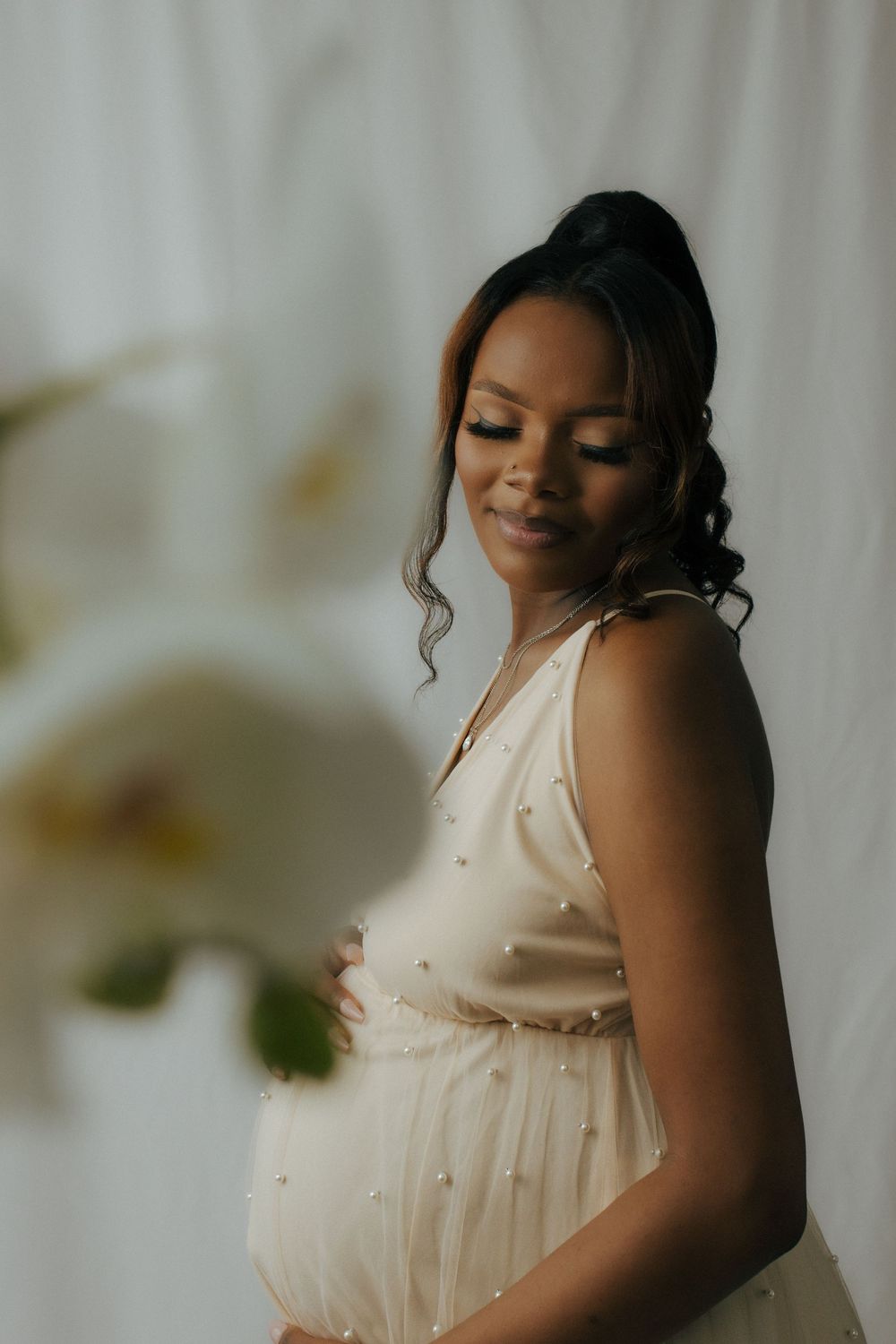 Expectant mother in cream-colored dress smiles serenely while cradling her baby bump in soft lighting.