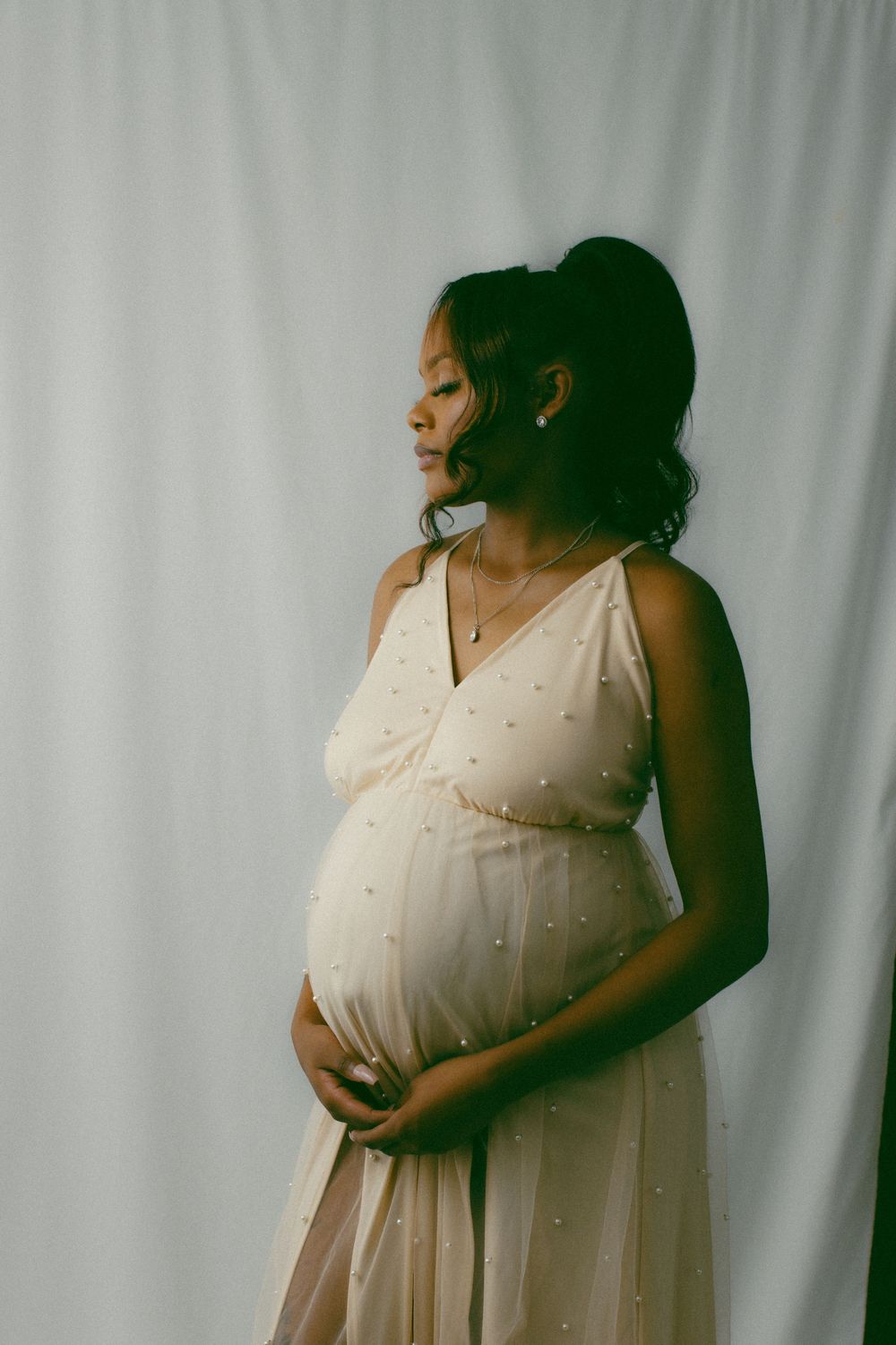 Side profile of an expectant mother in a white pearl-embellished dress standing against a light backdrop.