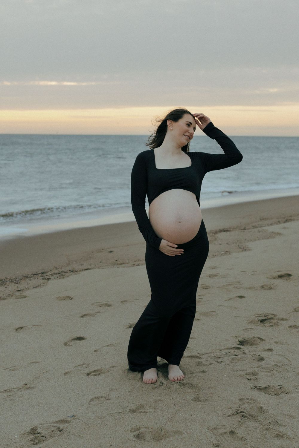 A pregnant woman in a black dress poses gracefully on a beach at sunset with the ocean in the background.