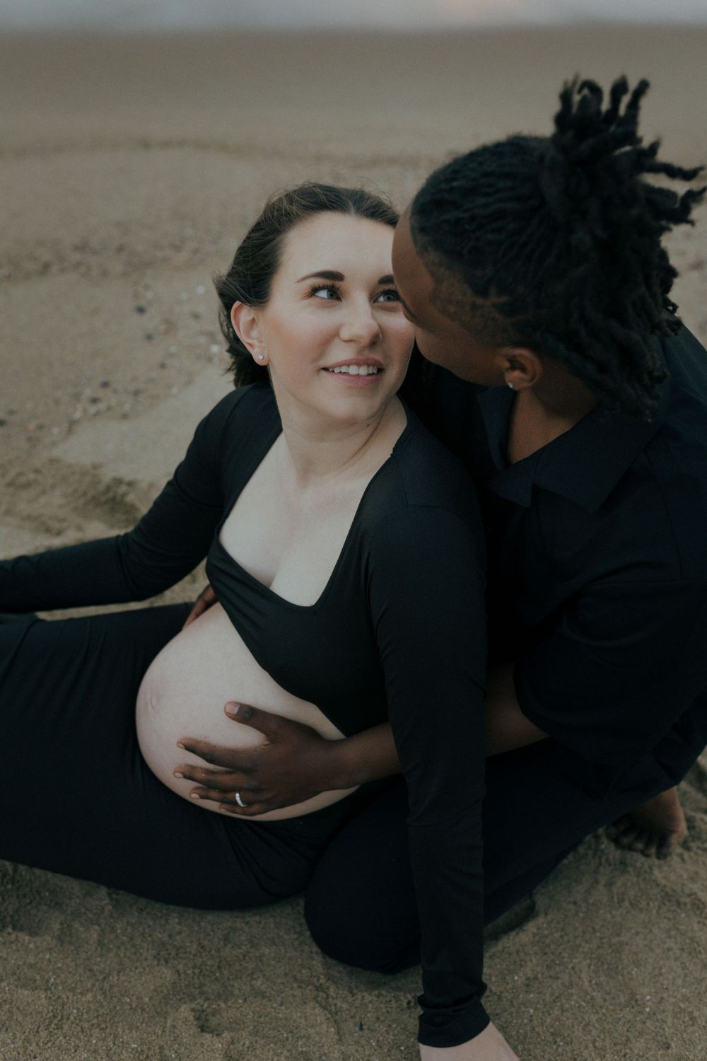 Intimate maternity photoshoot featuring a couple in black outfits embracing on sandy beach at dusk.