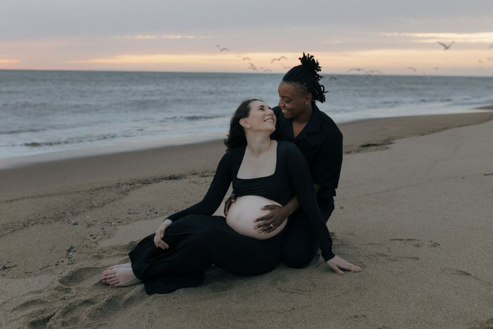 A couple embraces on the beach at sunset, sitting close together on the sand by the ocean.