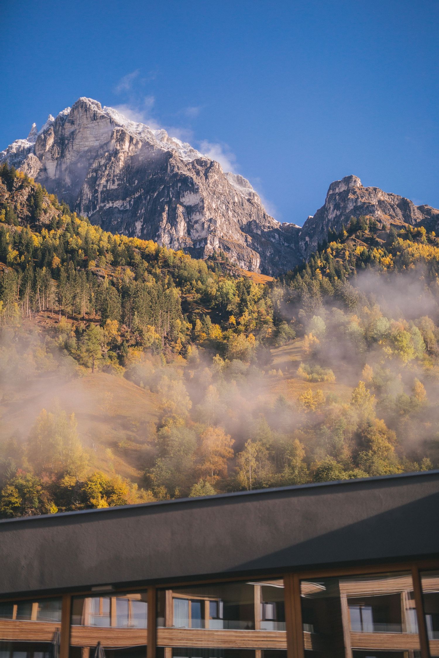 Majestic mountain peaks tower over a lodge with misty autumn foliage in golden colors against a blue sky.