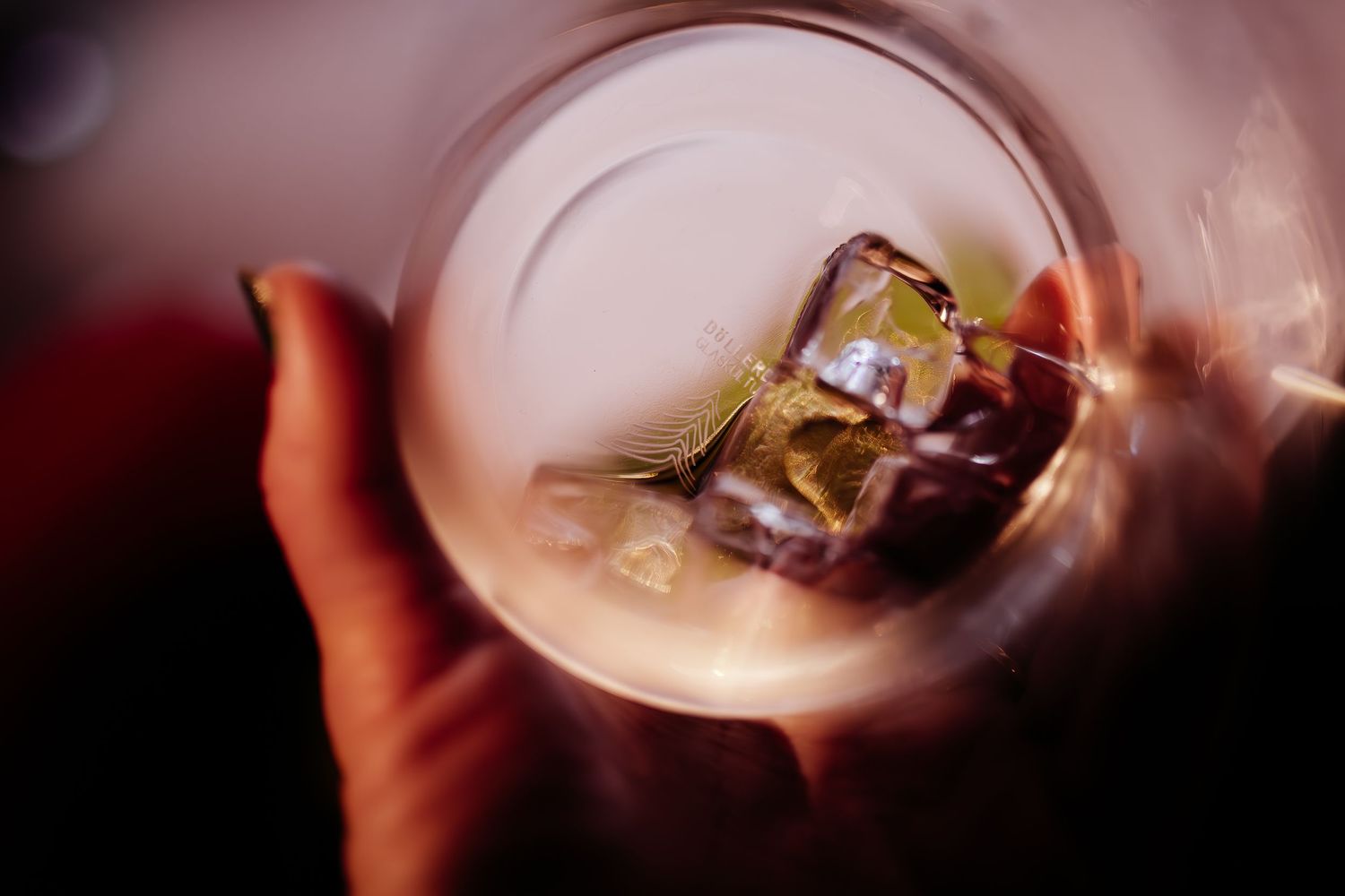 Glass of clear liquid with ice cubes viewed from above with hand holding it against dark background.