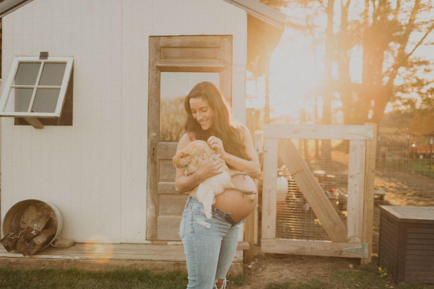 Person holds a small dog while standing outside a rustic white shed during golden hour.