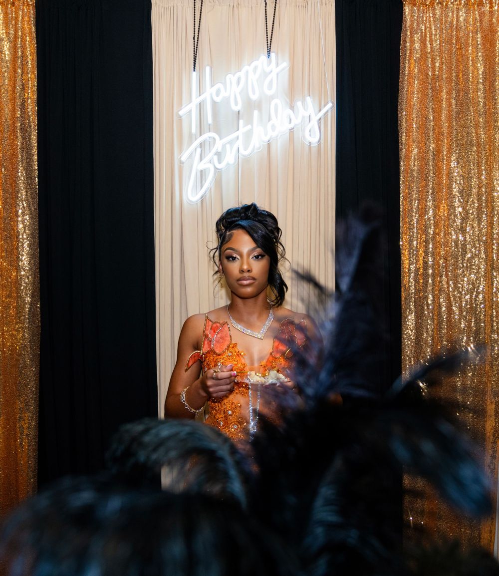 Woman stands before a Happy Birthday neon sign at a gold sequin backdrop celebration.