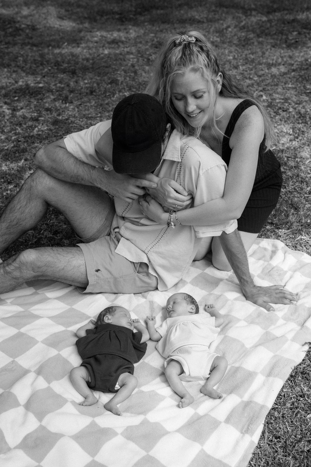 A couple sits together on a checkered blanket outdoors with twin babies lying beside them in black and white.
