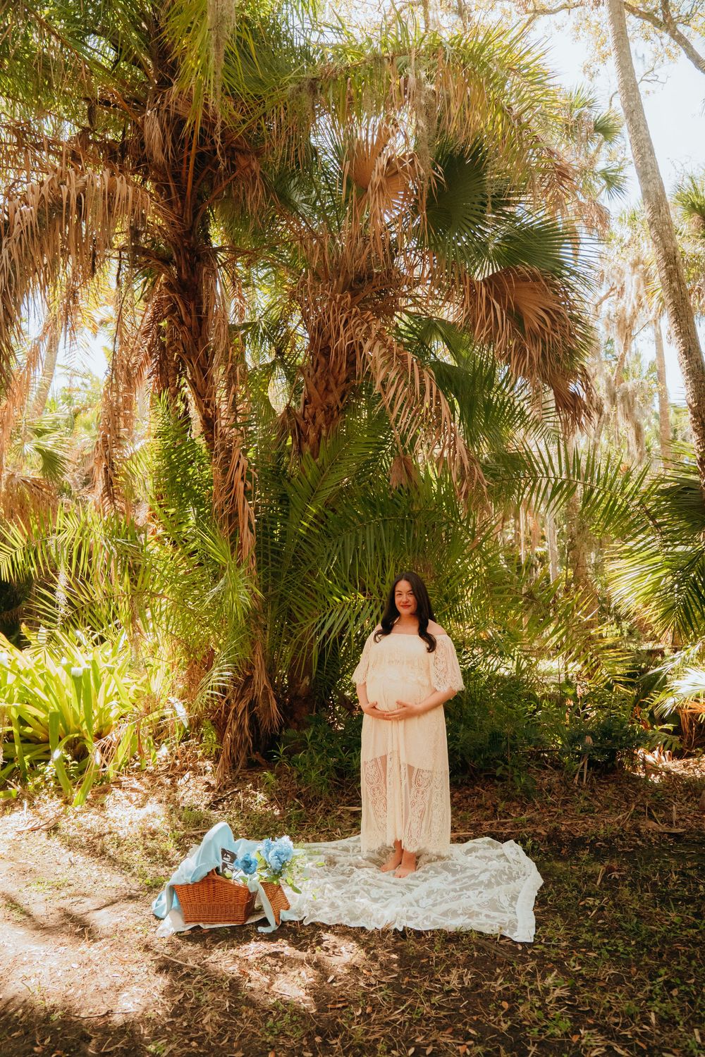 A person stands on a white blanket in a tropical garden setting with palm trees and natural sunlight.