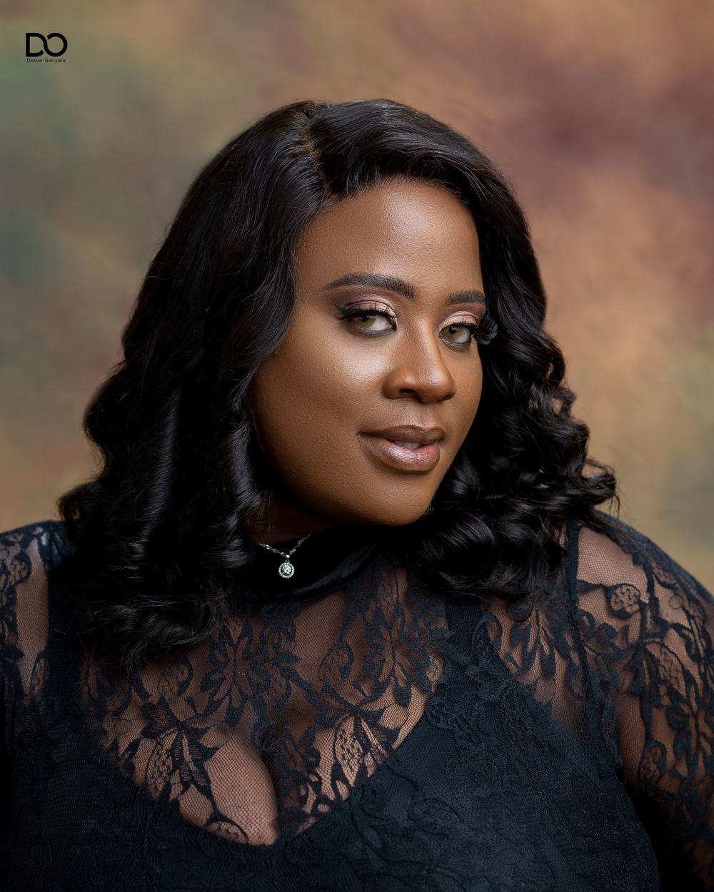 Professional portrait in black lace featuring elegant hair styling and jewelry against a soft brown backdrop.