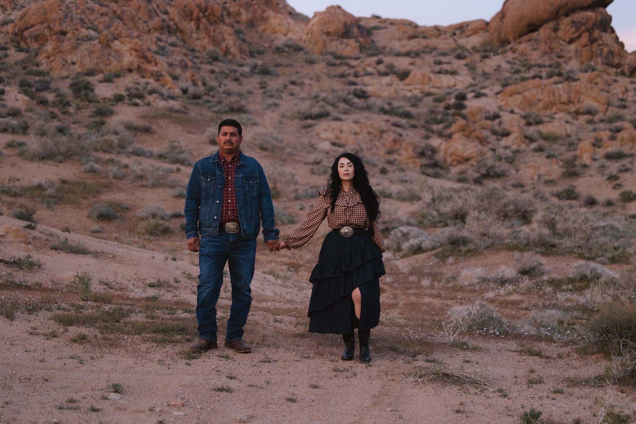Two people walk through a scenic desert landscape with red rock formations during sunset.