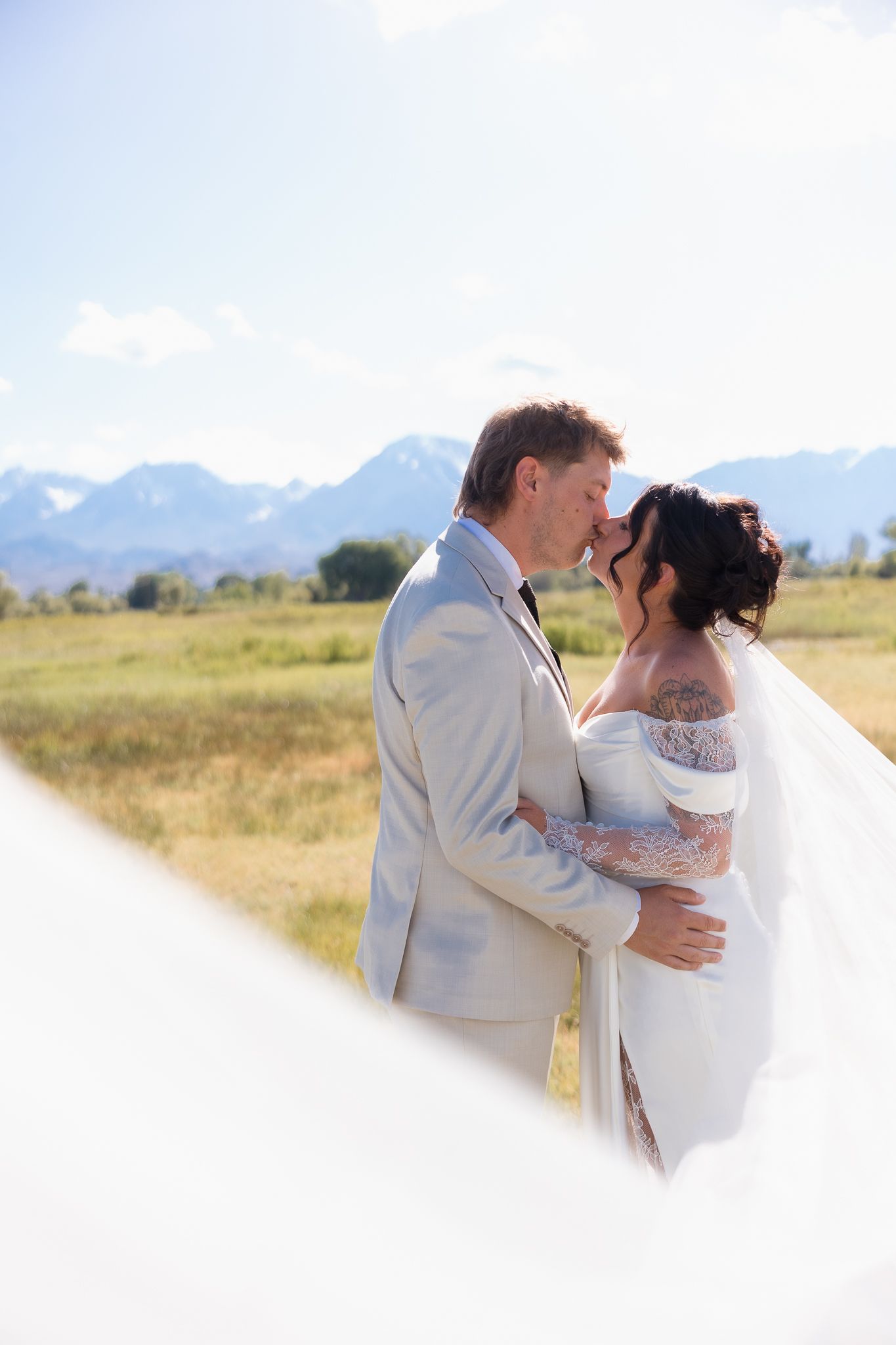 Bride and groom sharing a kiss in a sunlit mountain field, bride in lace off-shoulder gown with flowing veil.