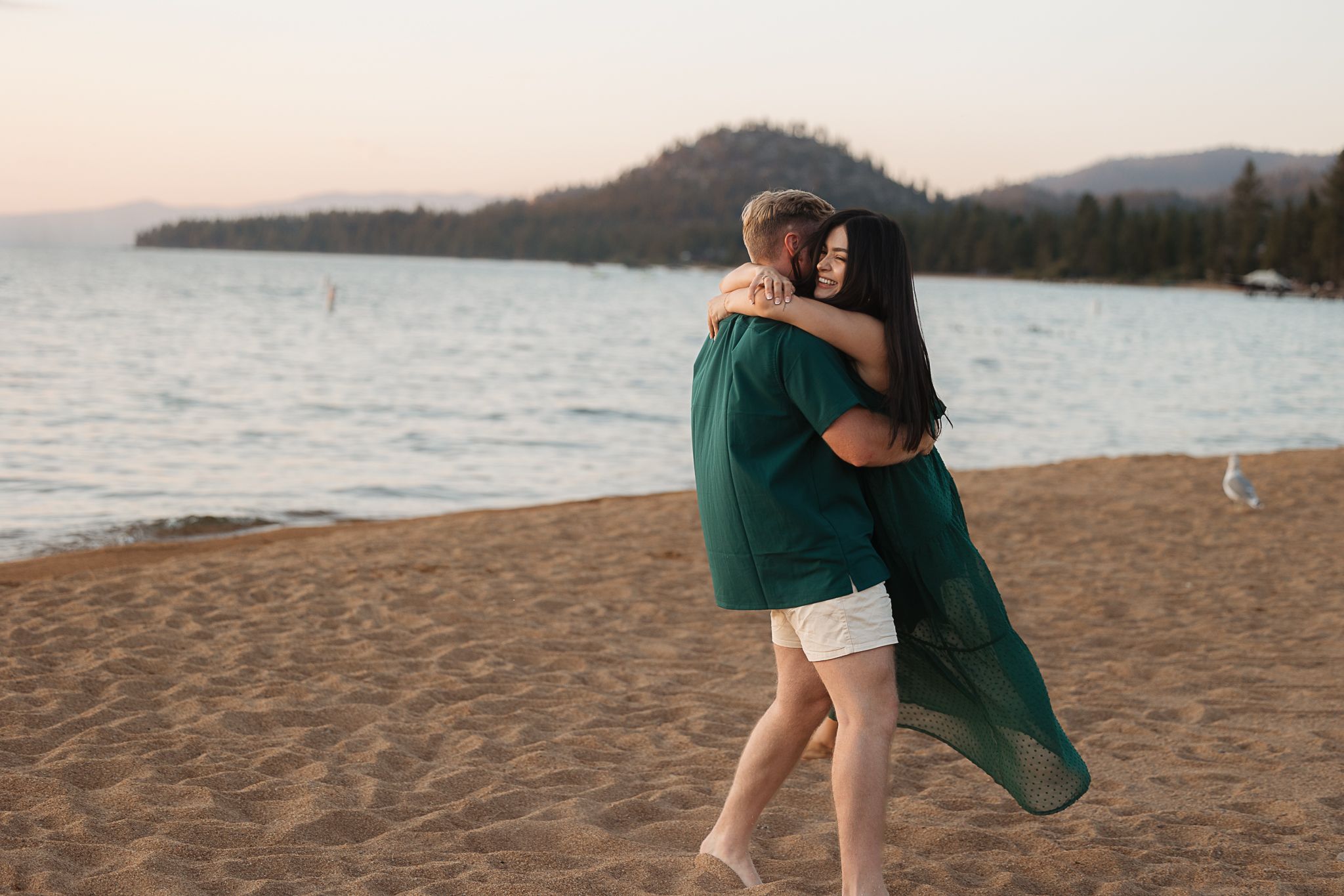 Couple shares romantic moments on a beach at sunset with mountains in background.