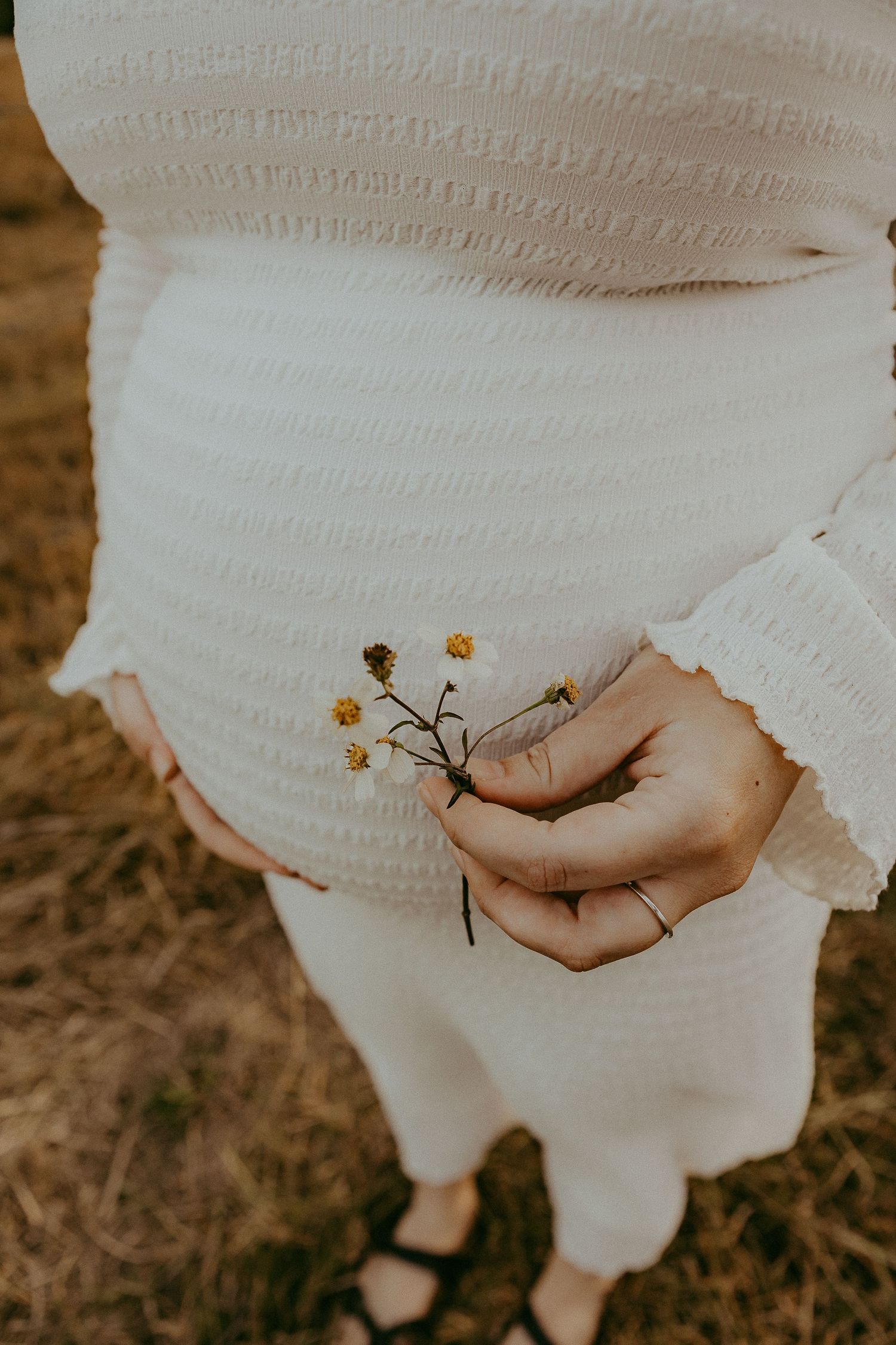 Close-up of pregnant woman in white dress holding tiny wildflowers against baby bump.