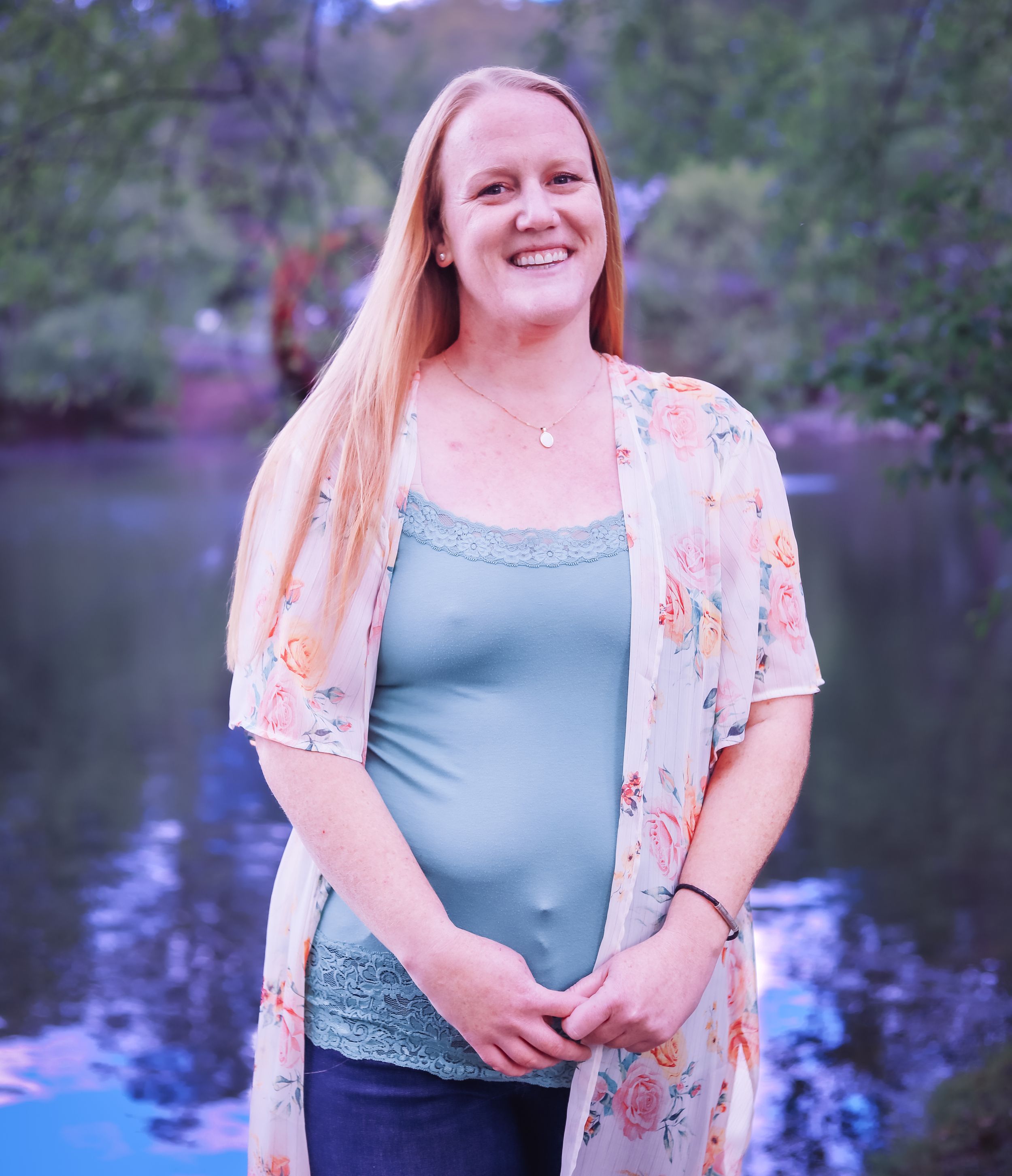 A person in a floral overlay and blue top poses near a lake with purple sunset tones.