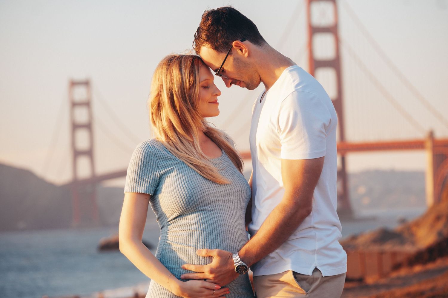 Intimate pregnancy photo shoot featuring a couple in white sweaters against a gray wall backdrop.