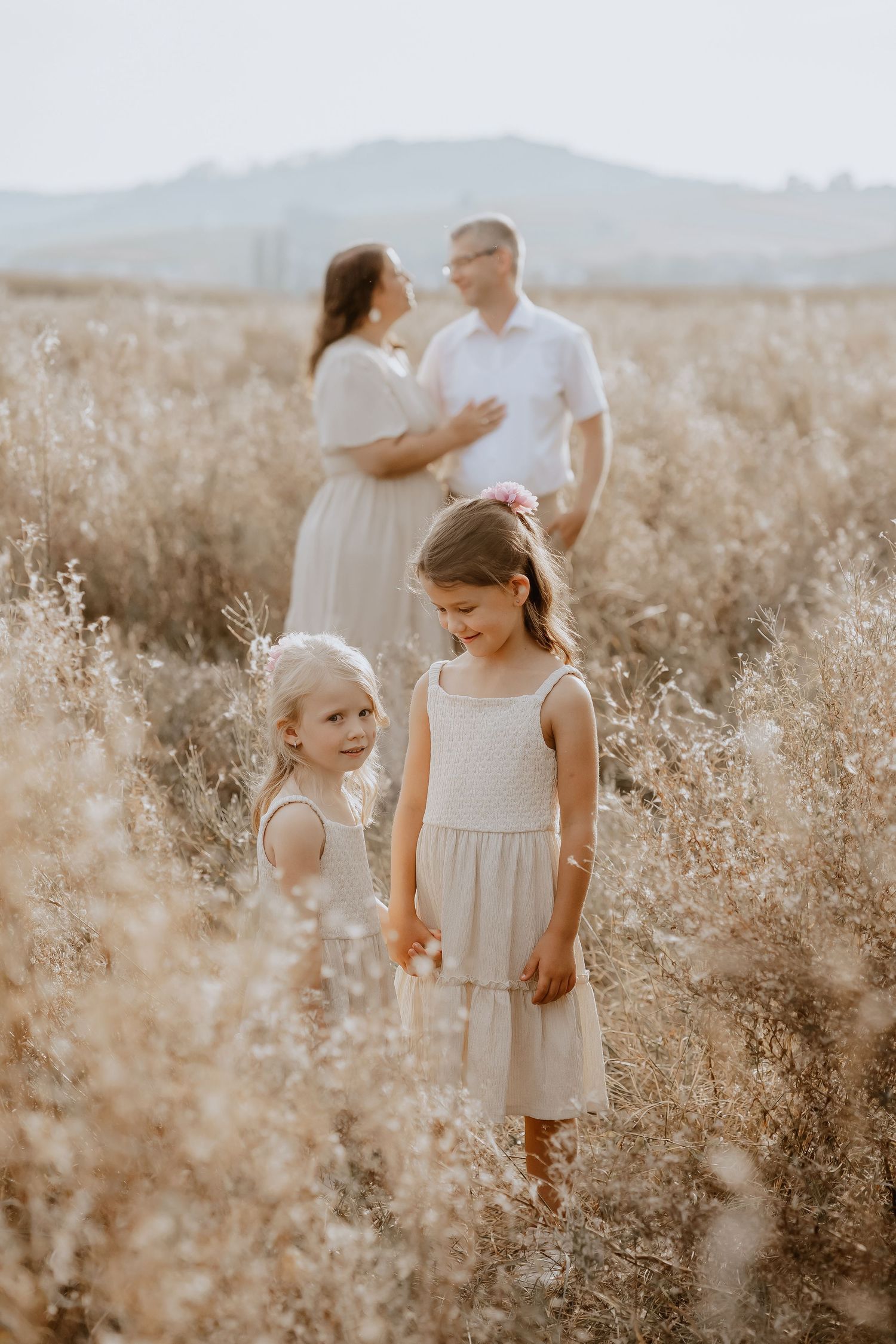 Parents in white clothing holding hands with their toddler walking through tall grass.