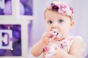 Baby eating cake while wearing pink floral headband during cake smash photoshoot with purple backdrop.