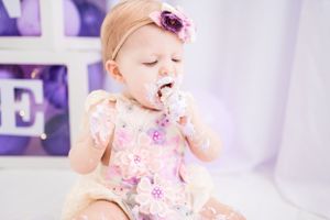 Baby enjoying cake during cake smash photo session with purple letter decorations in background.
