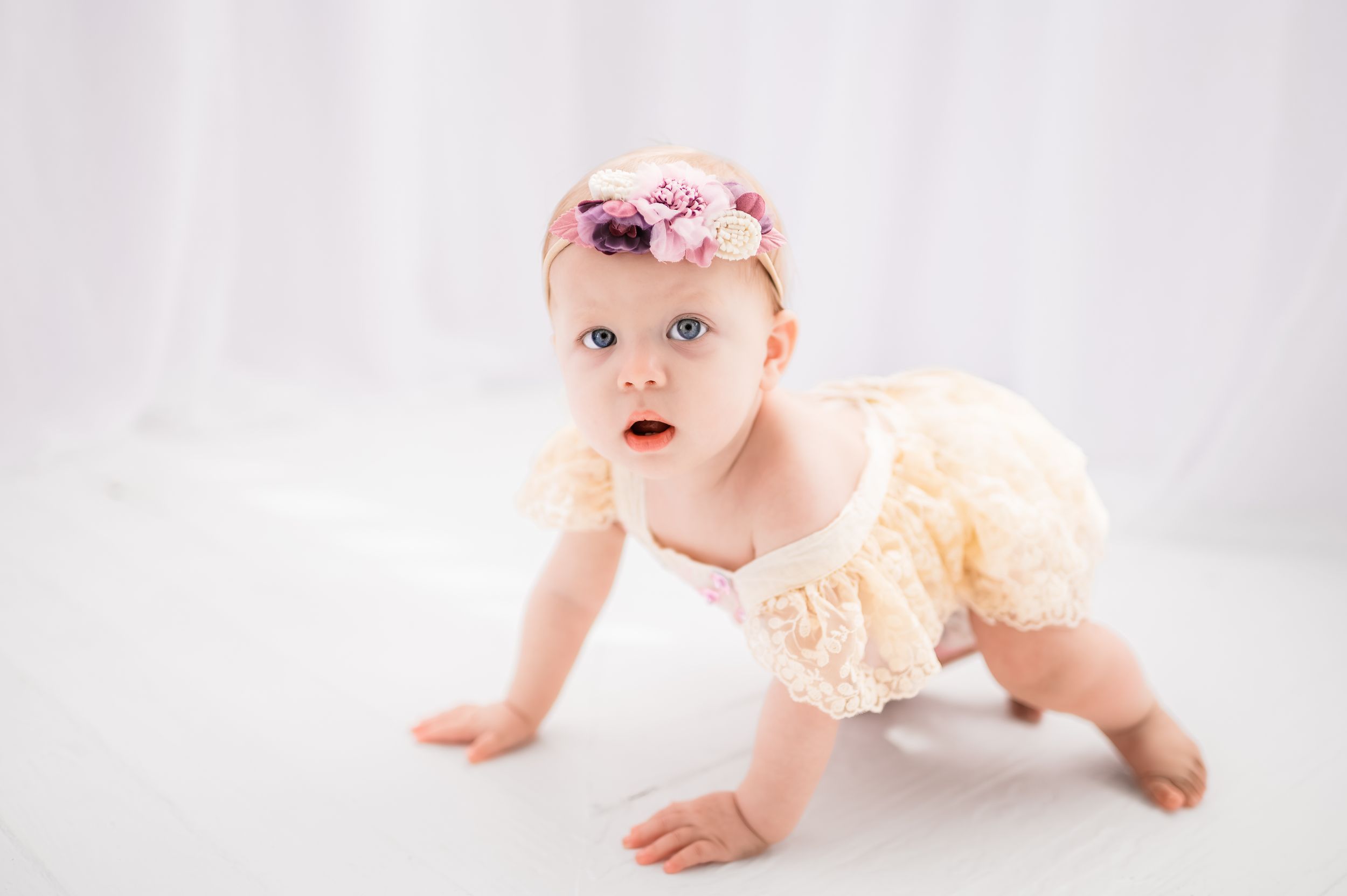 Baby in cream lace outfit crawling on white backdrop during portrait session.