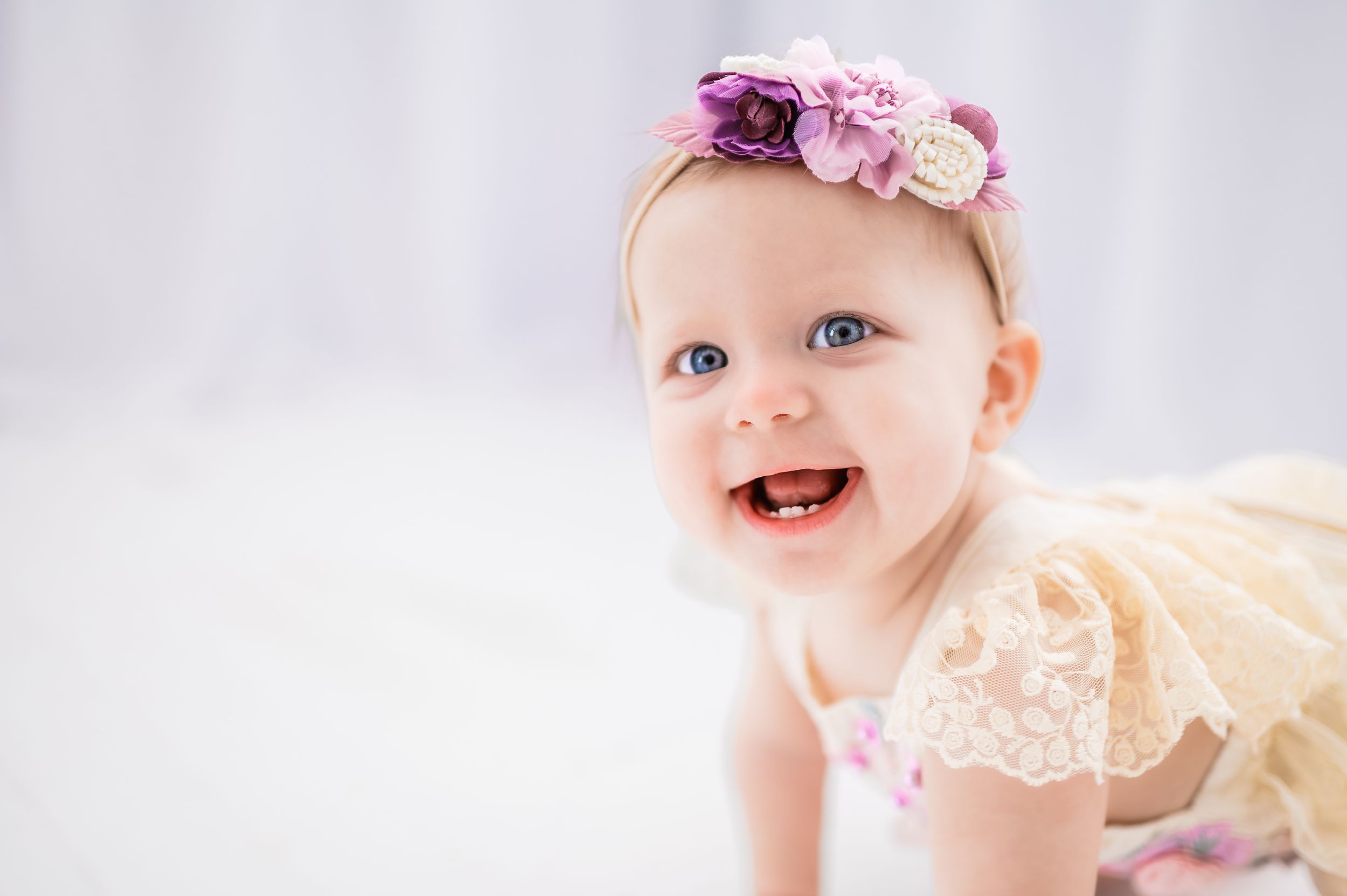 Baby wearing lace dress and floral headband smiles brightly against white background.