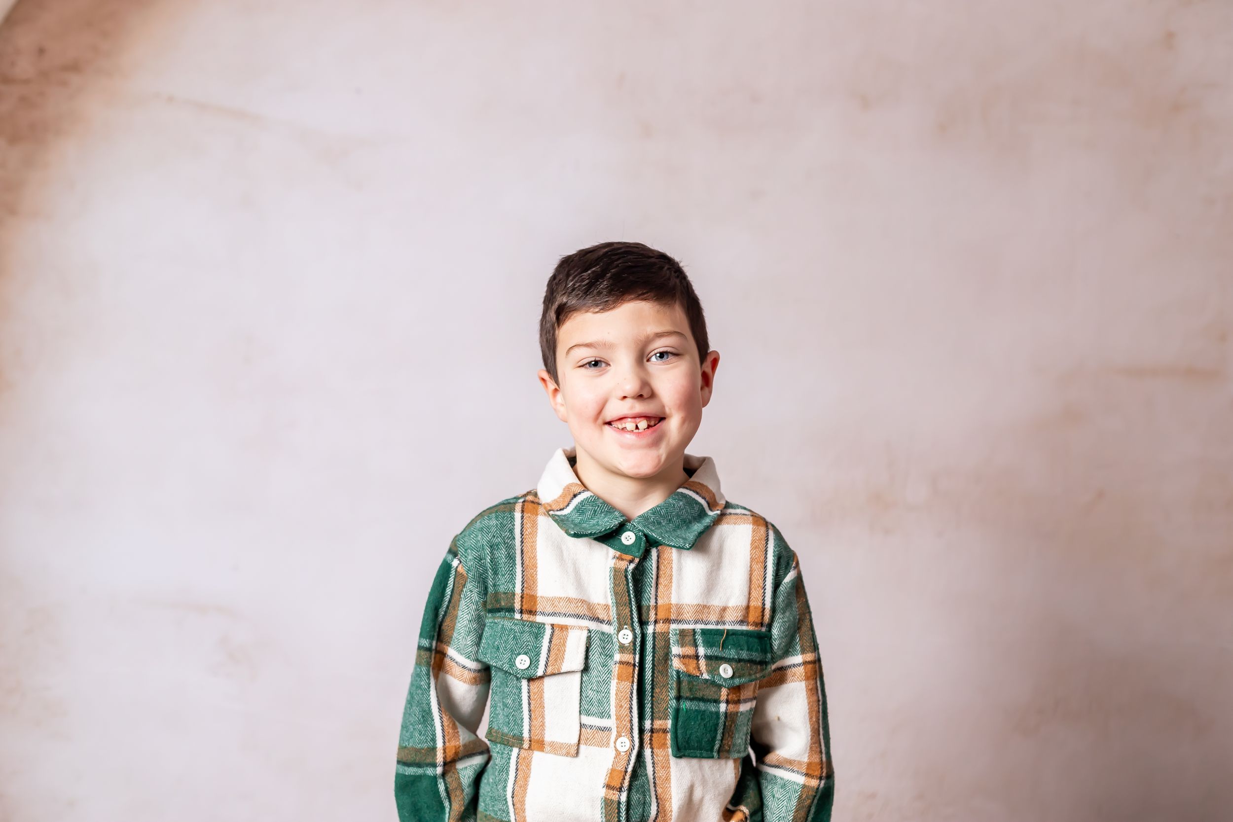 Close up portrait of child in plaid shirt smiling against beige backdrop.
