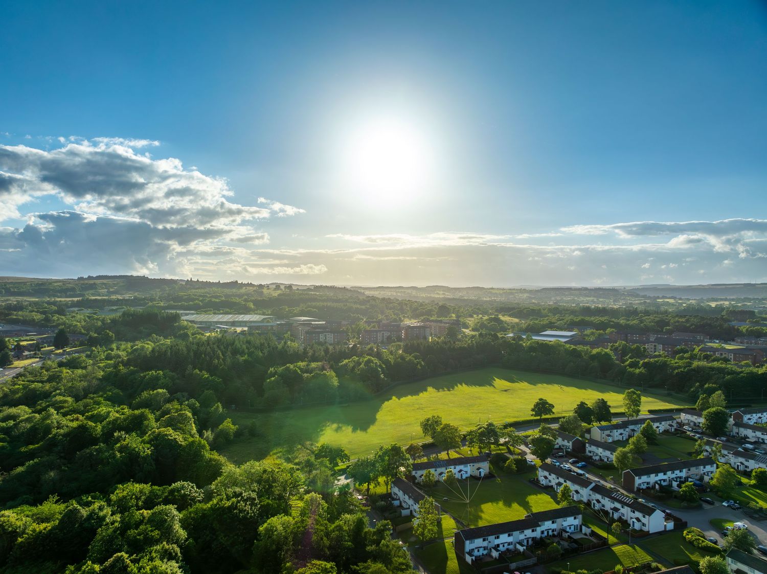 Aerial view of a sunlit suburban landscape with green fields and residential areas under blue sky.