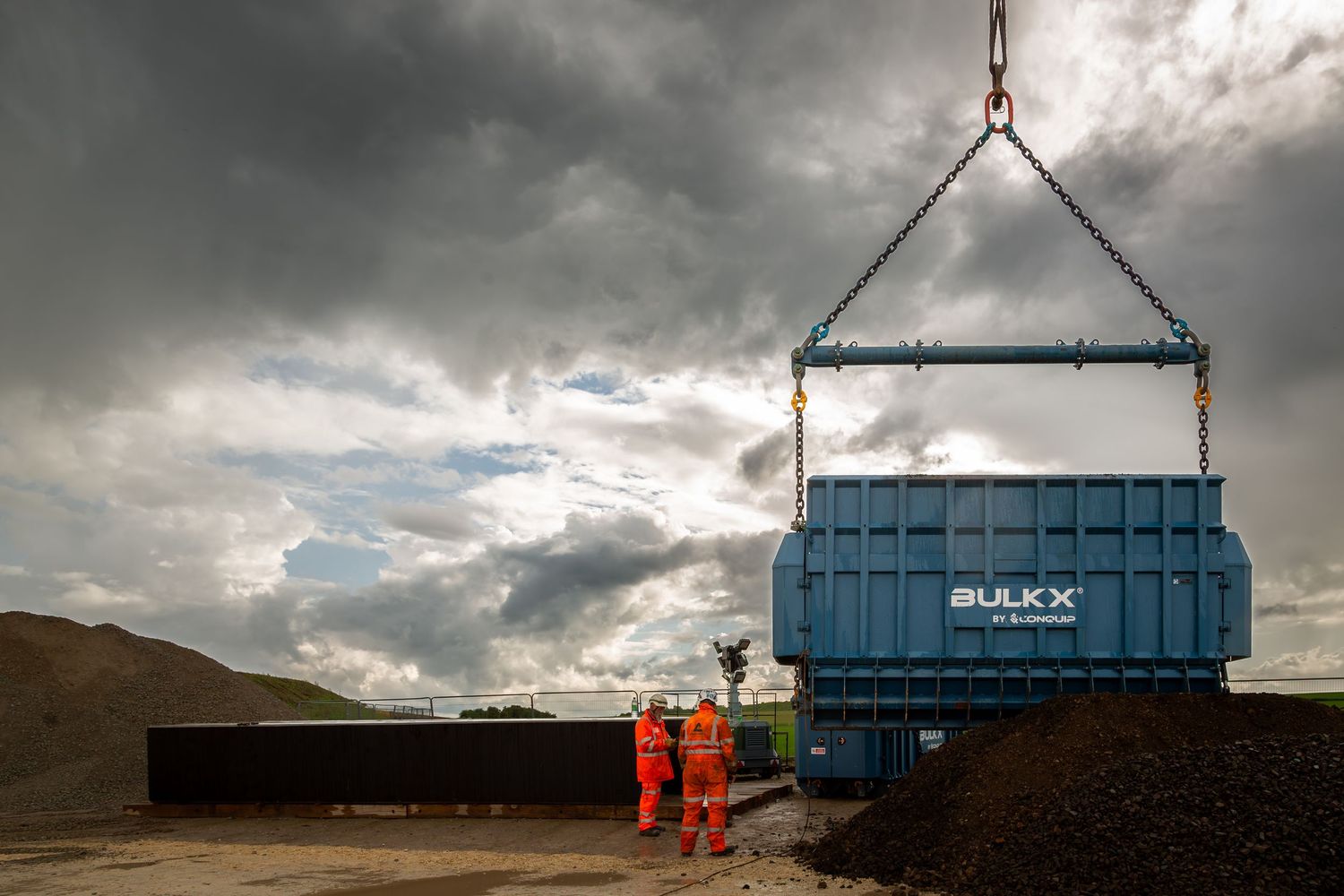 A series of shipping containers being lifted and installed on a construction site under dramatic cloudy skies.