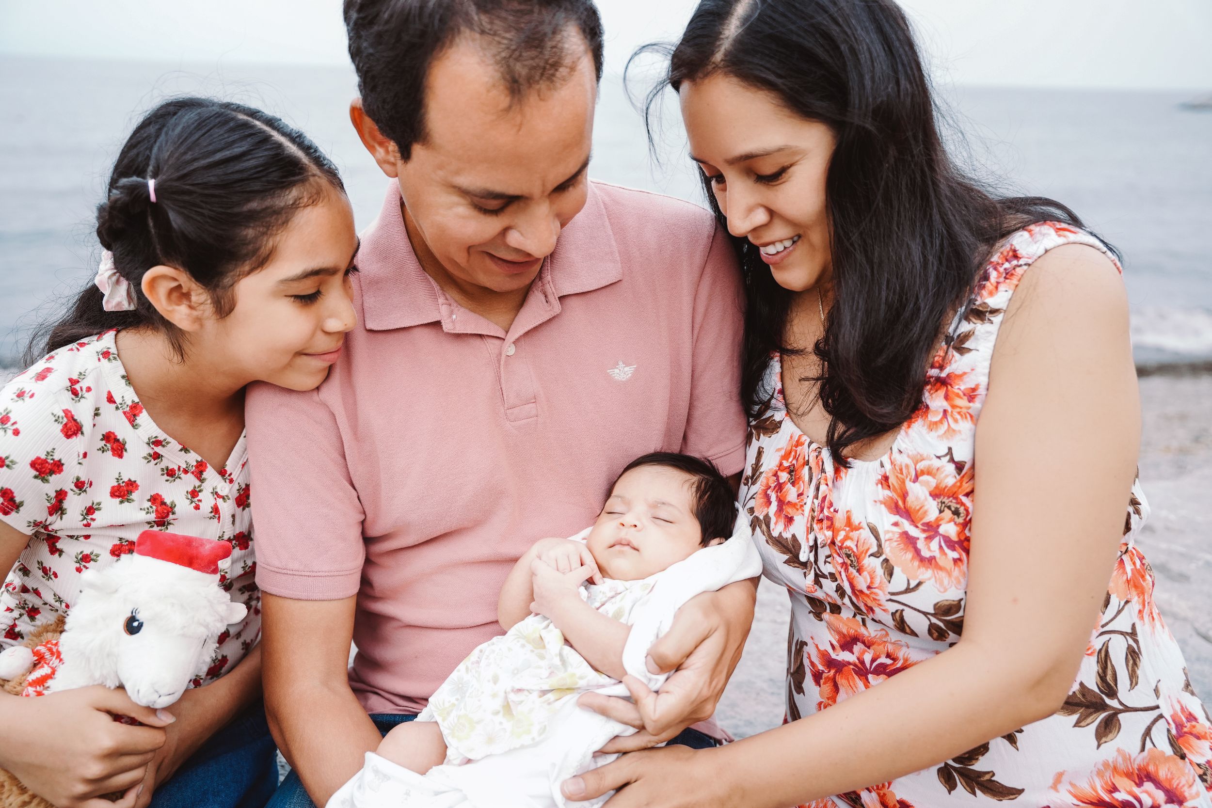 Family of four smiling at newborn baby on beach, parents with young daughter holding stuffed animal.