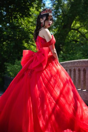 A woman in an elegant red ball gown with a large bow and crown poses gracefully in a lush green garden.