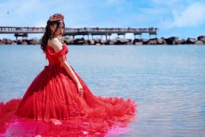 Woman in elegant red ballgown with crown poses gracefully by the ocean on a bright sunny day.