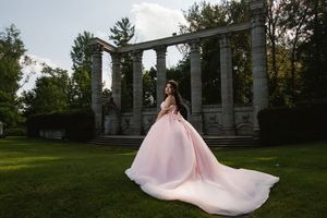 A woman in a flowing pink ball gown poses gracefully before stone columns in a lush green garden.